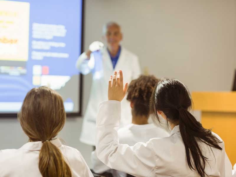 Clase de medicina con estudiantes en bata blanca, participando activamente durante una explicación práctica del profesor.
