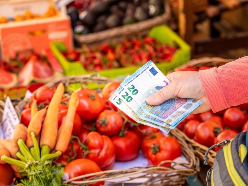 Mano pagando con billetes de euro por verduras frescas en un mercado, destacando el costo creciente de alimentos básicos por inflación.